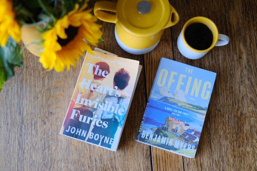 Two books on a coffee table with coffee cup and sunflowers in the background. The books are The Heart's Invisible Furies by John Boyne and The Offing by Benjamin Myers. 