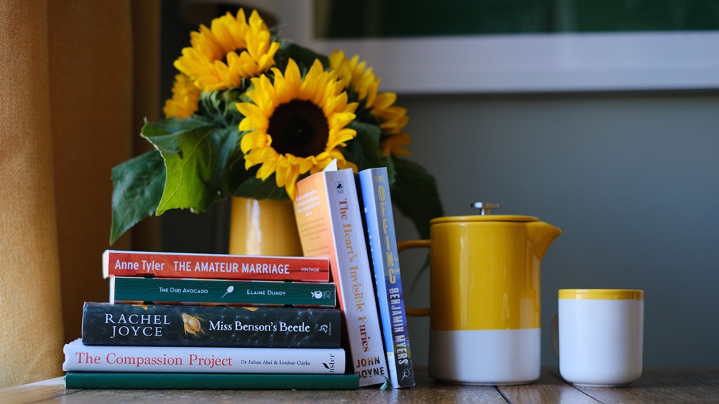 A table featuring a jug of sunflowers and a stack of books: The Amateur Marriage, The Dud Avocado, Miss Benson's Beetle, The Compassion Project, The Heart's Invisible Furies and The Offing