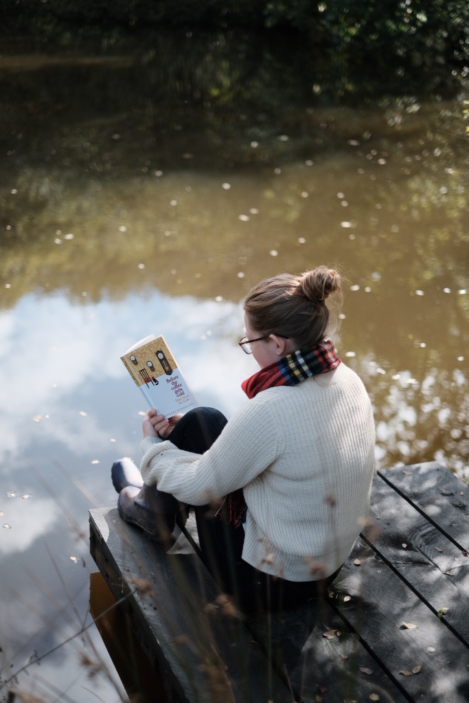 Libby sits on a deck by a lake reading Before the Coffee Gets Cold, wearing a jumper and scarf. 