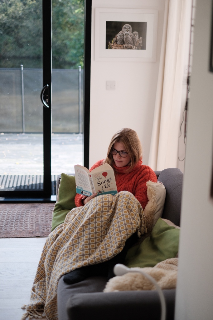 Libby sat on a sofa under a blanket and wearing an orange jumper, reading The Songs of Us. 